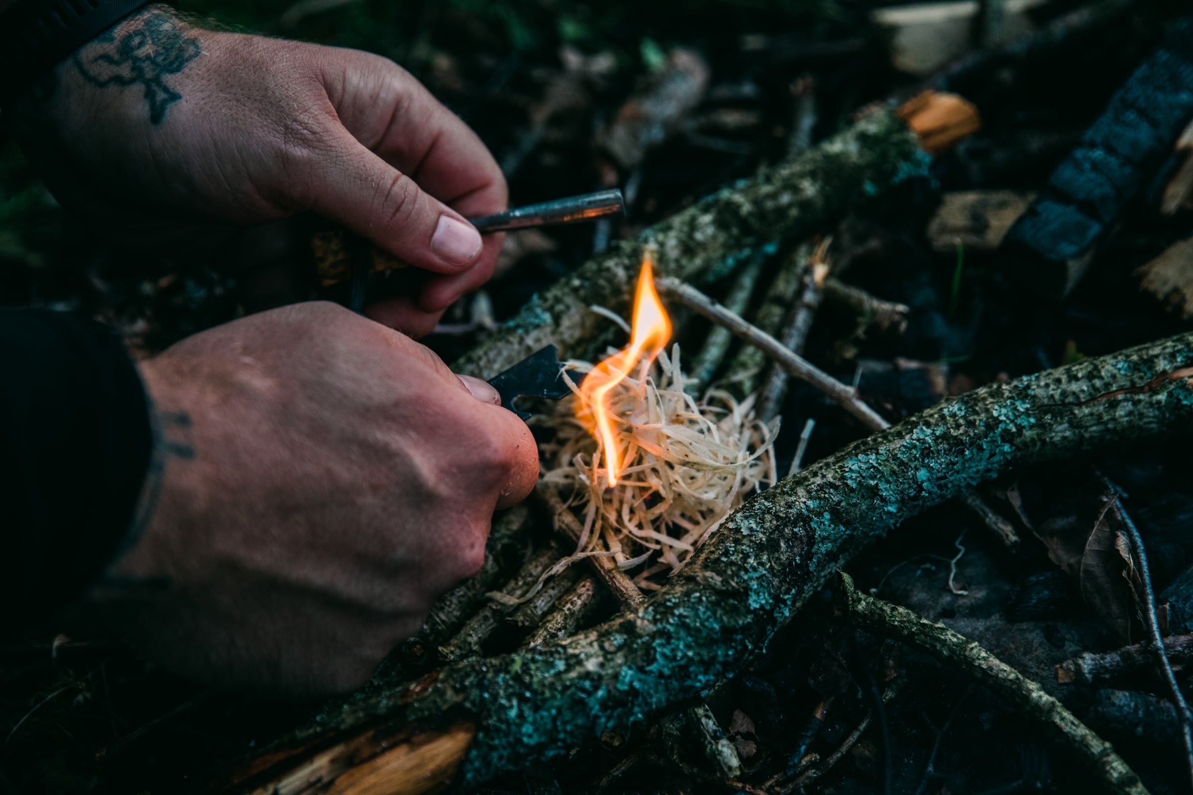 Millican | Close-up of hands using a flint to ignite a small fire on kindling, with a tattoo visible on one hand.