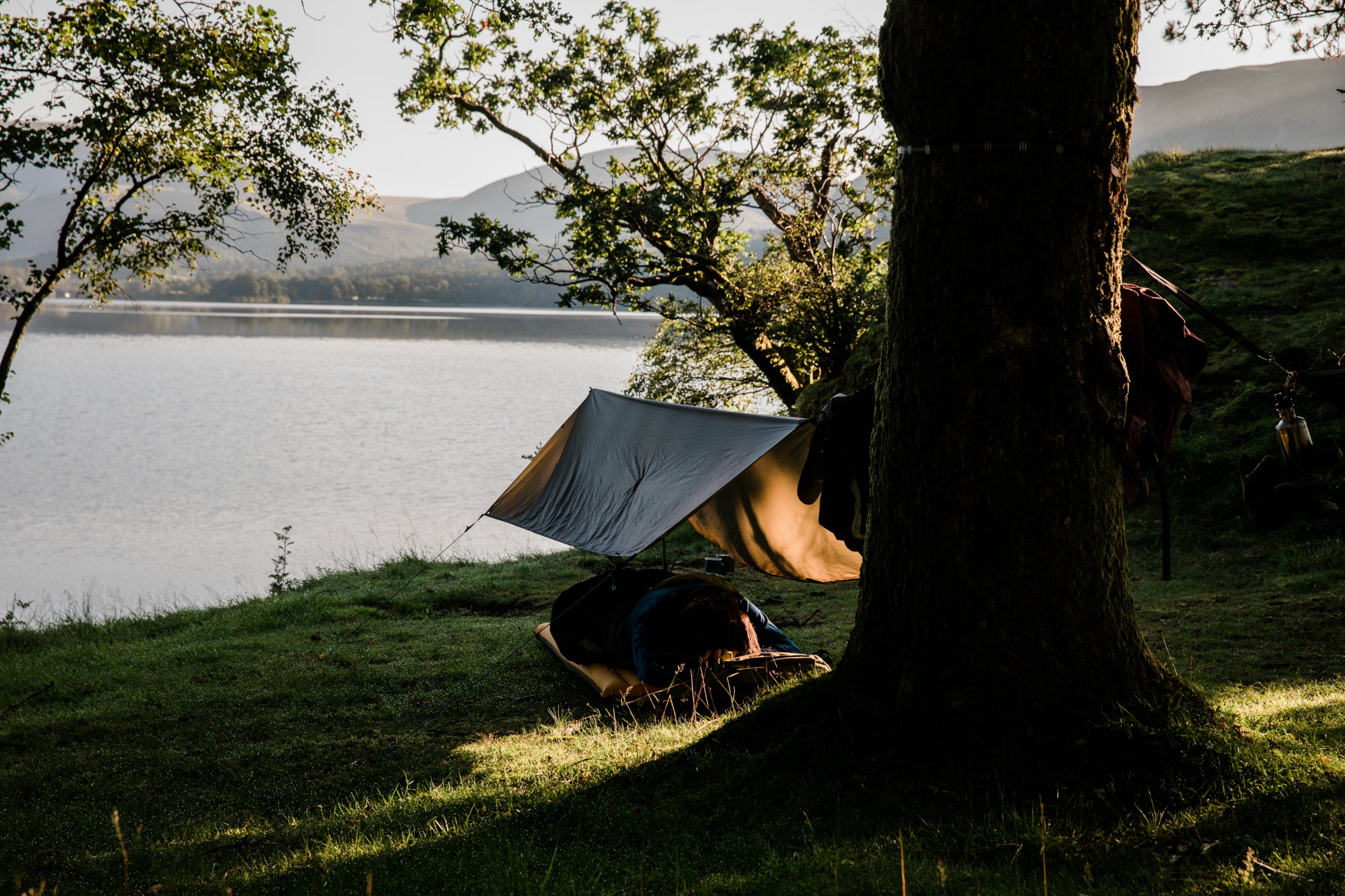 Millican | A hammock with a tarp hung between trees overlooking a tranquil lake at sunset, with clothes hanging on a line and outdoor gear scattered around, creating a serene camping scene.