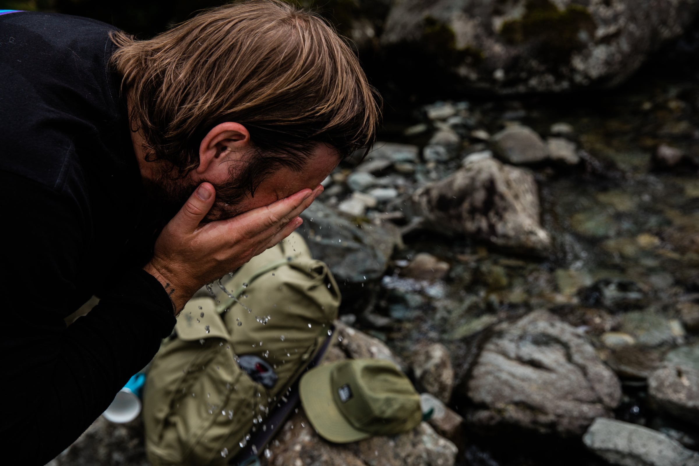 A man is washing his face with water from a rocky stream, with a green cap and Millican backpack placed beside him