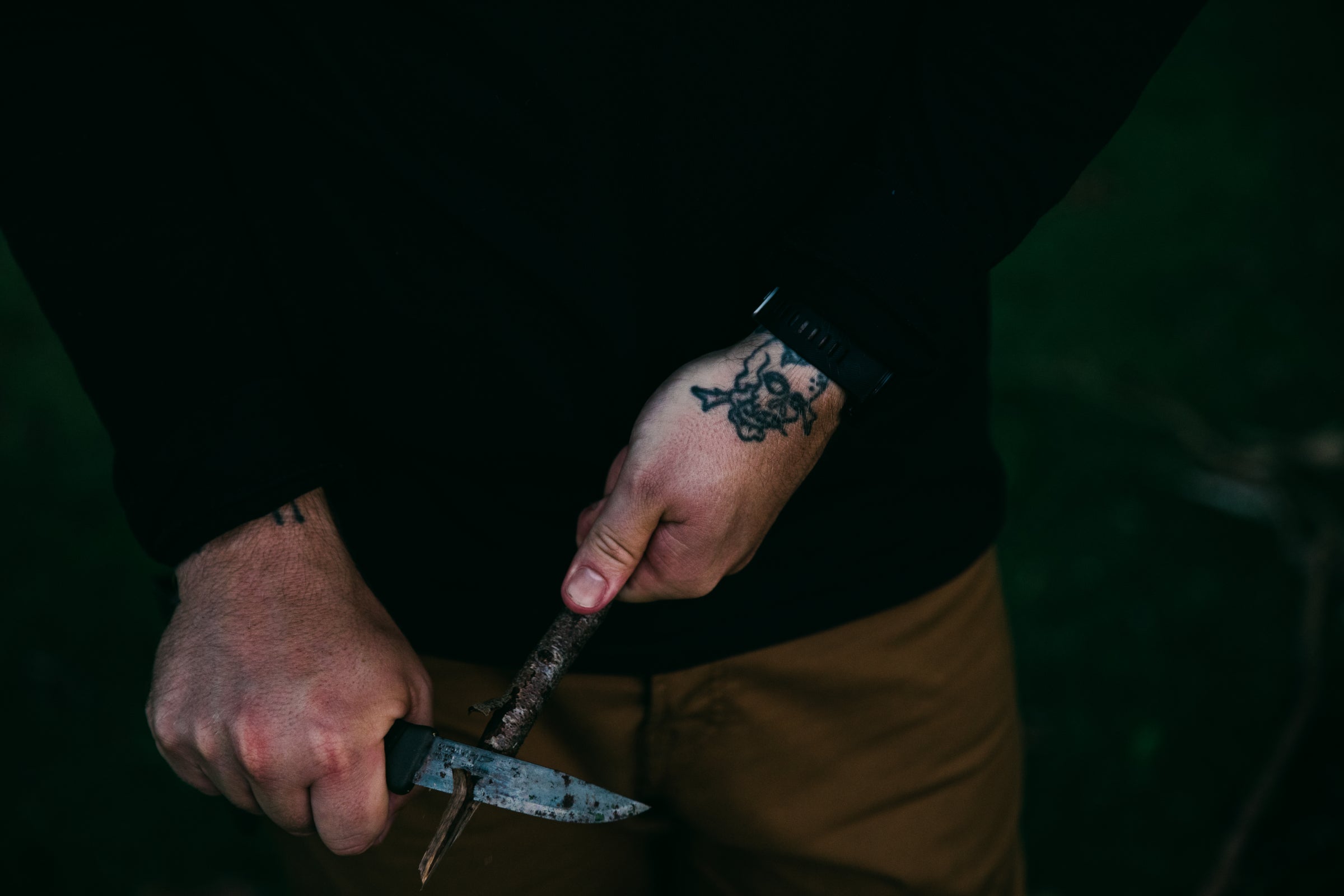 Millican | A person in dark clothing is carving a piece of wood with a knife. The individual's wrist displays a tattoo of a skull with a mustache, and they are wearing a black watch.